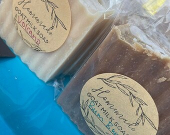 May include: Two bars of handmade goat milk soap wrapped in plastic. The soap on the left is labeled "Homemade Goat Milk Soap Volcano" and the soap on the right is labeled "Homemade Goat Milk Soap Rum  Rum".