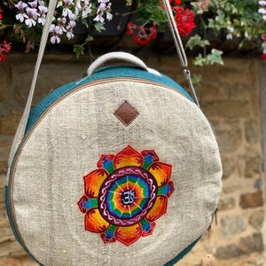 Peut inclure: Un sac rond bleu sarcelle et beige orné d'un motif de fleur de lotus brodé aux couleurs vives avec un symbole Om central. Le sac comprend une poignée supérieure, une bandoulière réglable et un petit écusson marron en forme de losange.