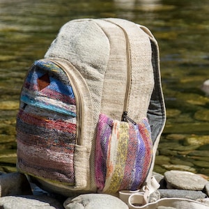 Op de afbeelding: Een beige rugzak met een gestructureerde, geweven stoffen body, voorzien van een voorvak en zijvak met horizontale strepen in levendige tinten blauw, rood, paars en geel. De tas bevat een klein bruin driehoekig detail als accent.