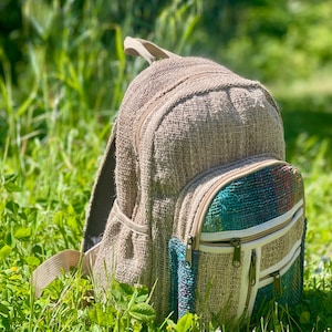 Pode incluir: Uma mochila de tecido bege com um compartimento principal grande e um bolso frontal menor com duas seções horizontais com zíper. O bolso frontal exibe um painel de tecido texturizado multicolorido em tons de azul, verde e vermelho, acentuado por um acabamento branco.