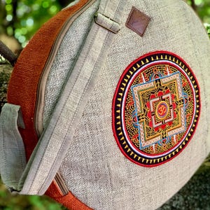 Op de afbeelding: Een ronde tas van beige en roestoranje stof met een verstelbare schouderband en ritssluiting. De voorzijde is voorzien van een cirkelvormige, veelkleurige geborduurde mandala-patch met een centraal Om-symbool en ingewikkelde geometrische patronen.