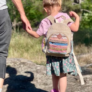 May include: A child wearing a small backpack with colourful woven details, a pink top, and a floral skirt. The child is holding hands with an adult and wearing pink sandals with bows.
