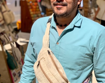 May include: A person wearing a light blue shirt and a gray baseball cap with a brown brim. They are wearing sunglasses and have a beard. They are wearing a beige hemp fanny pack with a zippered pocket and a colorful strap.