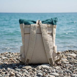 Pode incluir: Uma mochila de enrolar com corpo em lona bege natural e uma secção superior em tecido azul-petróleo escuro. A mochila inclui duas alças ajustáveis, um bolso horizontal com fecho no painel traseiro e bolsos laterais em ambos os lados.
