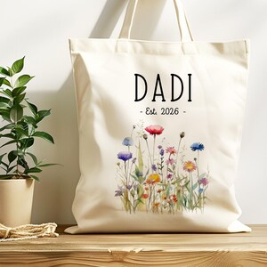 May include: Cream-colored tote bag with the word "DADI" in black, and "- Est. 2026 -" below. A watercolor floral design in pink, blue, purple, and yellow adorns the bottom. The bag has a simple, casual design.