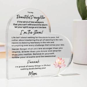 May include: A heart-shaped acrylic plaque with a message for a daughter. The message reads "To my Beautiful Daughter, If the wind of fate whispers that you can't withstand the storm, let your spirit surge and declare I'm the storm! Life isn't about waiting for the storm to pass, but rather about mastering the art of dancing in the rain. Here's to dancing fearlessly in the rain and triumphing over every challenge that comes your way. Never forget that you are stronger than you imagine, braver than you know and smarter than you realize. Believe in yourself, follow your dreams and be awesome! Hannah I'm proud of many things in life but nothing beats being your Mom."