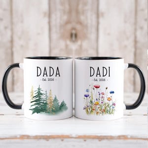 Op de afbeelding: Twee witte keramische mokken met zwarte handvatten en randen. Eén mok heeft het woord "DADA" boven "Est. 2026" en een aquarel boslandschap. De andere mok heeft "DADI" boven "Est. 2026" en een bloemenontwerp.