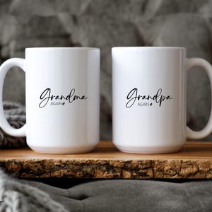 Op de afbeelding: Twee witte keramische mokken met zwarte tekst die "Grandma Again" en "Grandpa Again" luidt. Elke mok heeft een hartsymbool na "Again". De mokken staan op een houten oppervlak.