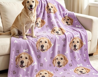 May include: A light purple fleece blanket featuring a repeating pattern of golden retriever dog faces, white bone shapes, and grey paw prints. A golden retriever dog sits on the blanket, which is draped over a light-colored sofa.