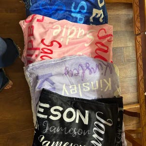 K&ouml;nnte beinhalten: Ein Stapel aus vier weichen Pl&uuml;schdecken in Blau, Rosa, Lavendel und Schwarz. Jede Decke zeigt ein sich wiederholendes Muster eines bestimmten Namens in verschiedenen Schriftarten und Gr&ouml;&szlig;en, darunter Sadie, Kinsley und Jameson in heller Schrift.