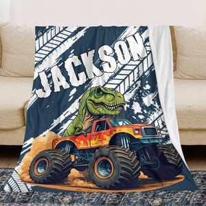 May include: A personalized blanket featuring a monster truck with a green dinosaur on top. The truck is orange and yellow, kicking up dirt. The name "JACKSON" is printed in large white letters against a navy blue background.