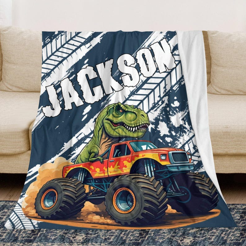 May include: A personalized blanket featuring a monster truck with a green dinosaur on top. The truck is orange and yellow, kicking up dirt. The name "JACKSON" is printed in large white letters against a navy blue background.