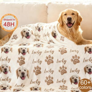 May include: A cream-colored blanket featuring a repeating pattern of golden retriever faces, paw prints, and the word "Lucky" in brown. A golden retriever dog is lying on the blanket. The blanket is made in the USA.