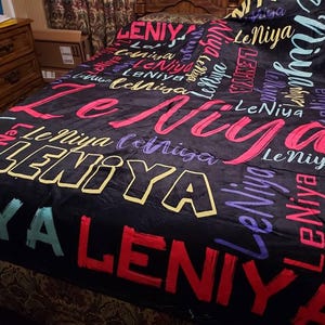 K&ouml;nnte beinhalten: Eine schwarze Fleecedecke mit einem sich wiederholenden Muster des Namens LeNiya in verschiedenen Schriftarten, Gr&ouml;&szlig;en und Farben, darunter Pink, Gelb, Lila, Rot und T&uuml;rkis. Der Text ist in Schreib- und Blockschrift &uuml;ber die Fl&auml;che verteilt.