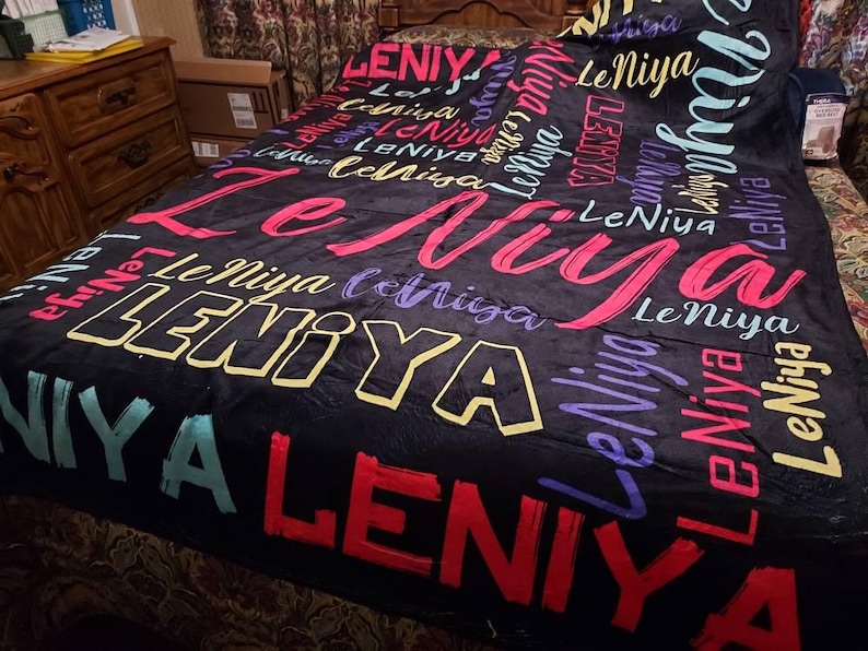 K&ouml;nnte beinhalten: Eine schwarze Fleecedecke mit einem sich wiederholenden Muster des Namens LeNiya in verschiedenen Schriftarten, Gr&ouml;&szlig;en und Farben, darunter Pink, Gelb, Lila, Rot und T&uuml;rkis. Der Text ist in Schreib- und Blockschrift &uuml;ber die Fl&auml;che verteilt.