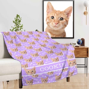 May include: A lilac-colored blanket featuring a repeating pattern of orange tabby cat faces and white fish bones. The word "COOKIE" is printed in white on a lilac band. A framed portrait of an orange tabby cat hangs on the wall.