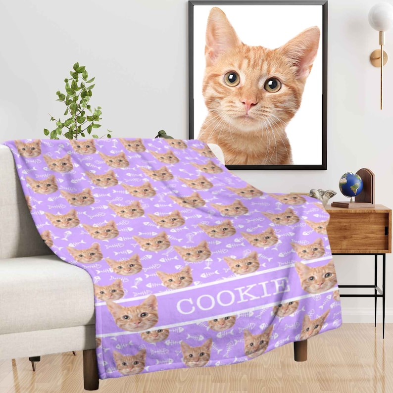 May include: A lilac-colored blanket featuring a repeating pattern of orange tabby cat faces and white fish bones. The word "COOKIE" is printed in white on a lilac band. A framed portrait of an orange tabby cat hangs on the wall.