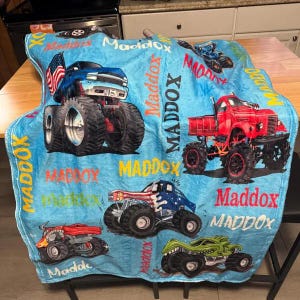 May include: A light blue blanket featuring illustrations of monster trucks in red, blue, and green. The blanket has the name "Maddox" printed in multiple places. The monster trucks have large tires and patriotic designs.