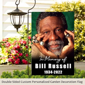May include: A black and green garden flag with a photo of Bill Russell, a basketball player, wearing championship rings. The text "In Memory of Bill Russell 1934-2022" is printed on the flag.