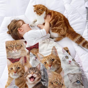 May include: A white blanket with a collage of different cats printed on it. The cats are all different colors and have different expressions. Some of the cats are wearing glasses or bow ties. A young person is lying on the blanket with a cat in their arms.