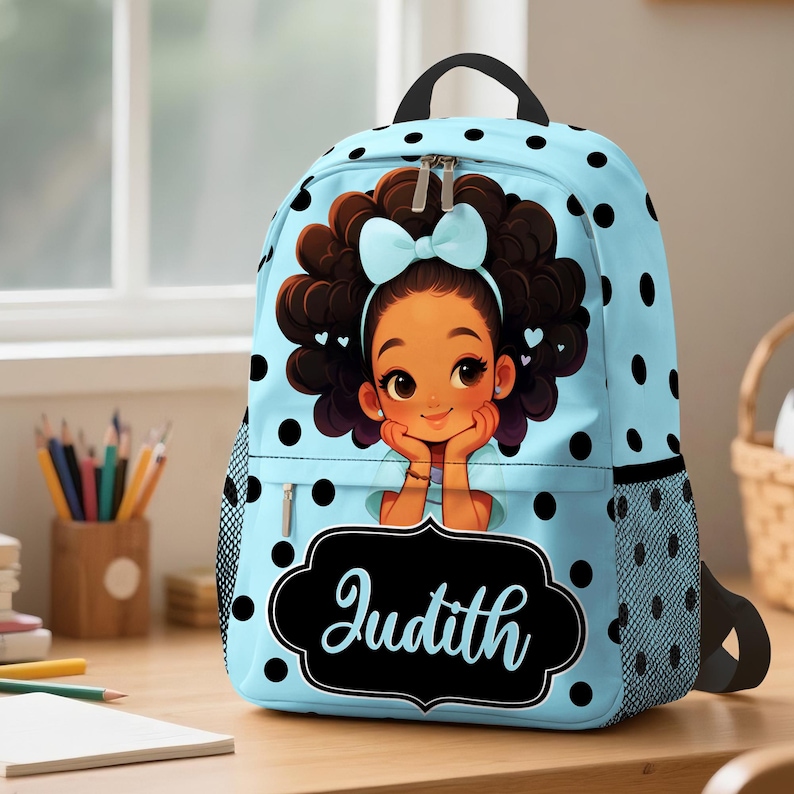 May include: A light blue backpack featuring a black polka dot pattern and a graphic illustration of a young person with dark curly hair and a light blue bow. The front pocket displays a black decorative label with the name Judith in white.
