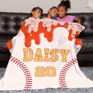 Op de afbeelding: Een witte deken met een honkbalthema, met rode stiksels, de naam "DAISY" in wafelletters en het nummer "20". De bovenkant van de deken heeft een ontwerp dat lijkt op een dessert met oranje glazuur en hagelslag.