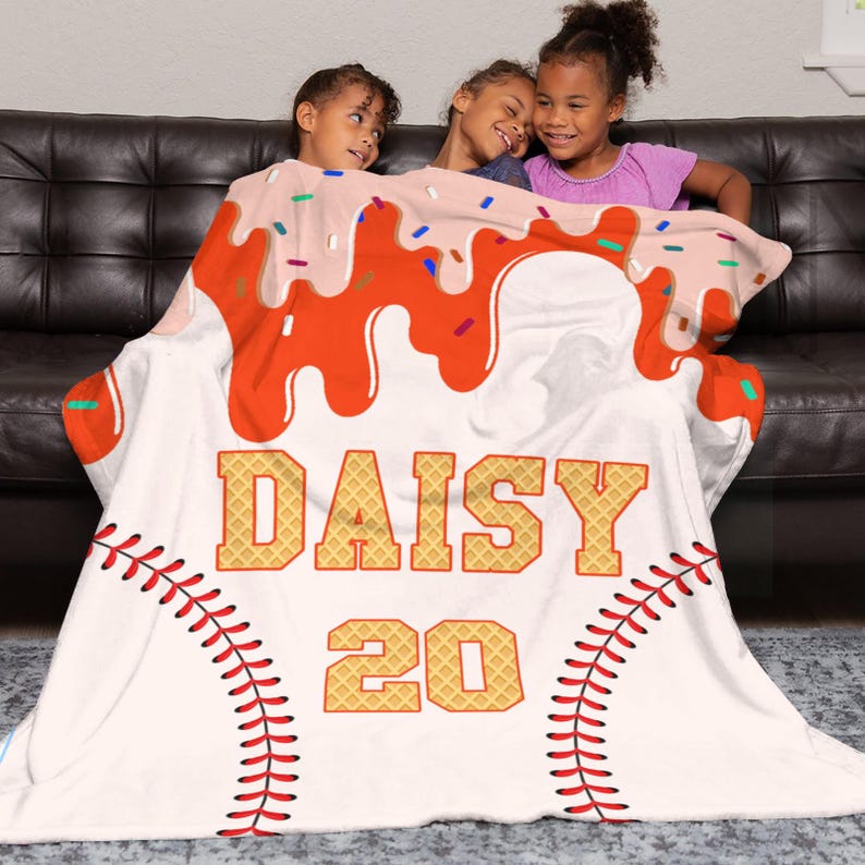 Op de afbeelding: Een witte deken met een honkbalthema, met rode stiksels, de naam "DAISY" in wafelletters en het nummer "20". De bovenkant van de deken heeft een ontwerp dat lijkt op een dessert met oranje glazuur en hagelslag.