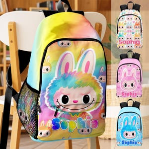 May include: A colorful backpack with a rainbow gradient design, featuring a cartoon bunny character with large eyes and a smiling face. The name "Sophia" is printed in a playful font. Additional backpacks in pink, blue, and white are also shown.