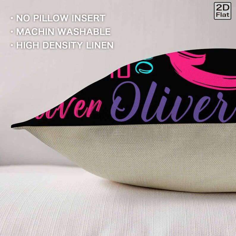 K&ouml;nnte beinhalten: Ein dekorativer Kissenbezug aus hochdichtem Leinen mit einem schwarzen oberen Bereich, der den Text Oliver in violetter Schreibschrift und rosa Akzenten zeigt. Der untere Bereich ist einfarbig beige. Der Text auf dem Bild lautet NO PILLOW INSERT, MACHIN WASHABLE und HIGH DENSITY LINEN.