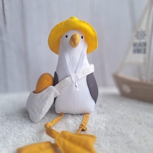 May include: A soft seagull toy, featuring a yellow hat, beak, and webbed feet. The seagull wears a white sailor collar and carries a small orange object in a white pouch. It has black wing accents and is designed to sit upright.