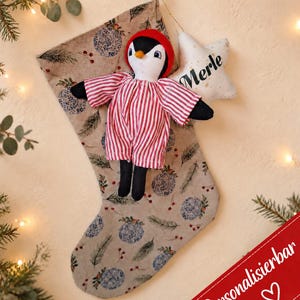 Könnte beinhalten: Ein beiger Weihnachtsstrumpf mit einem Muster aus blauen Ornamenten und Tannennadeln, verziert mit einer Plüsch-Pinguinpuppe in rot-weiß gestreiftem Schlafanzug und einem weißen sternförmigen Anhänger mit dem Namen Merle in schwarzer Schrift.