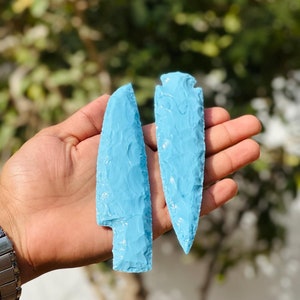 May include: Two light blue, hand-crafted flint arrowheads. One arrowhead is a knife shape, and the other is a classic arrowhead shape. The arrowheads are held in a person's hand, with a blurred green background.