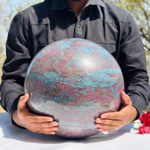 May include: A large, polished sphere with a swirling pattern of teal, burgundy, and grey. The stone is held by a person, showcasing its size and intricate colour variations. The sphere appears to be made of a natural stone.
