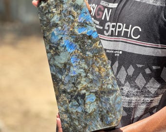 May include: A large, rectangular labradorite crystal with a polished surface. The stone displays a mix of dark gray, blue, and iridescent flashes. The crystal is held in both hands, showcasing its size and natural beauty.