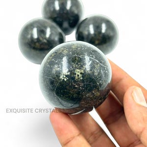 Op de afbeelding: Vier donkere, bolvormige kristallen met een gepolijst, reflecterend oppervlak. De kristallen hebben een gespikkelde uitstraling met hints van goud en groen. De tekst "EXQUISITE CRYSTALS" is zichtbaar. De kristallen worden in een hand gehouden.