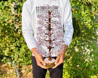 Könnte beinhalten: Eine Person hält einen violetten Amethyst-Kristallbaum mit einer Holzbasis. Der Baum besteht aus Draht und Kristallen und ist ein beliebtes Dekorationsobjekt.