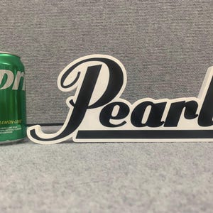 May include: White and black plastic sign with the word "Pearl" in a stylized font. The sign is on a gray surface.