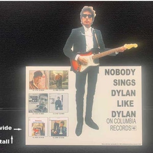 May include: A white poster with a black and white image of Bob Dylan playing a guitar. The poster reads "Nobody Sings Dylan Like Dylan on Columbia Records". The poster is 12 inches wide and 14 inches tall.