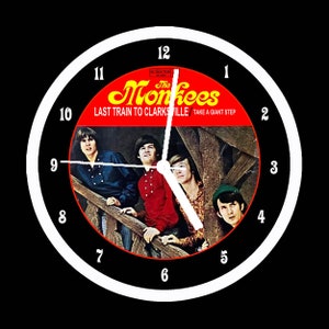 May include: Round wall clock featuring the album cover of The Monkees' "Last Train to Clarksville." The clock face has a black background with white numerals and hands. The album cover shows the band members in a vintage style.