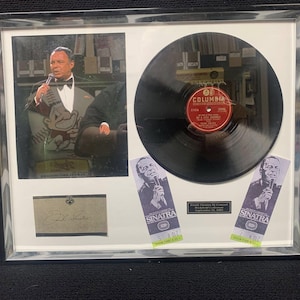May include: Framed memorabilia featuring a photograph of Frank Sinatra, a vinyl record, and two concert tickets. The record has a red and gold label. The frame is silver and contains a signed card.