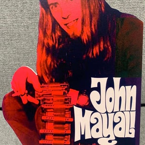 May include: A vintage advertisement featuring John Mayall, a musician, playing a Hohner Blues Harp harmonica. The image is in red, white, and blue colors with a retro design.