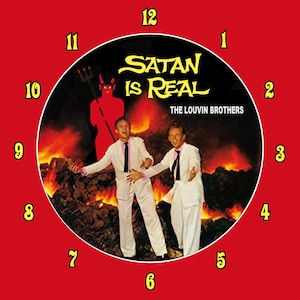 May include: A clock face with a black background and a red border. The clock face features a photo of two men in white suits standing in front of a fiery background. The text "SATAN IS REAL" is written in yellow letters above the men. The text "THE LOUVIN BROTHERS" is written in white letters below the men. The clock face has yellow numbers around the edge.