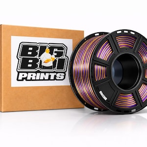 May include: A brown cardboard box with a white label featuring the text "BIG BOI PRINTS" and a cartoon figure. Next to it is a black spool of purple and gold filament for 3D printing.