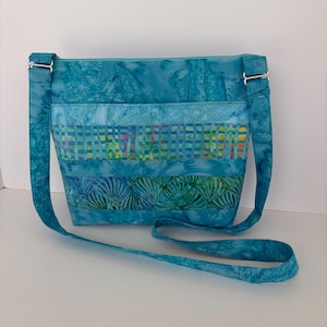 May include: A turquoise crossbody bag with a patterned design. The bag features a long, adjustable strap and silver-colored hardware. The bag's design incorporates various shades of blue and green, with a textured, batik-style pattern.