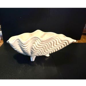 May include: Cream-colored ceramic clam shell bowl with a wavy, textured surface. The bowl is supported by three small feet. The design mimics the natural form of a clam shell, suitable for decorative or functional use.