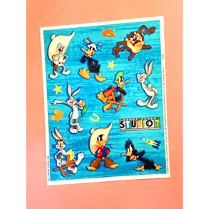 May include: A sheet of colorful stickers featuring characters from the Looney Tunes cartoon series. The stickers include Bugs Bunny, Daffy Duck, Tweety Bird, and Sylvester the Cat. The stickers are on a blue background with white stars and the words "Looney Tunes Studio" in a banner.