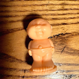 May include: A small, peach-colored plastic doll with a round face, chubby cheeks, and a smiling expression. The doll is wearing a diaper and has a foot with the text "©1984 OAA".