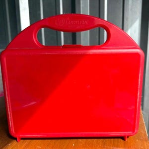 May include: A bright red plastic lunchbox with a handle and the text "SANDY LION" on the handle. The lunchbox is rectangular with rounded corners and sits on a wooden surface.