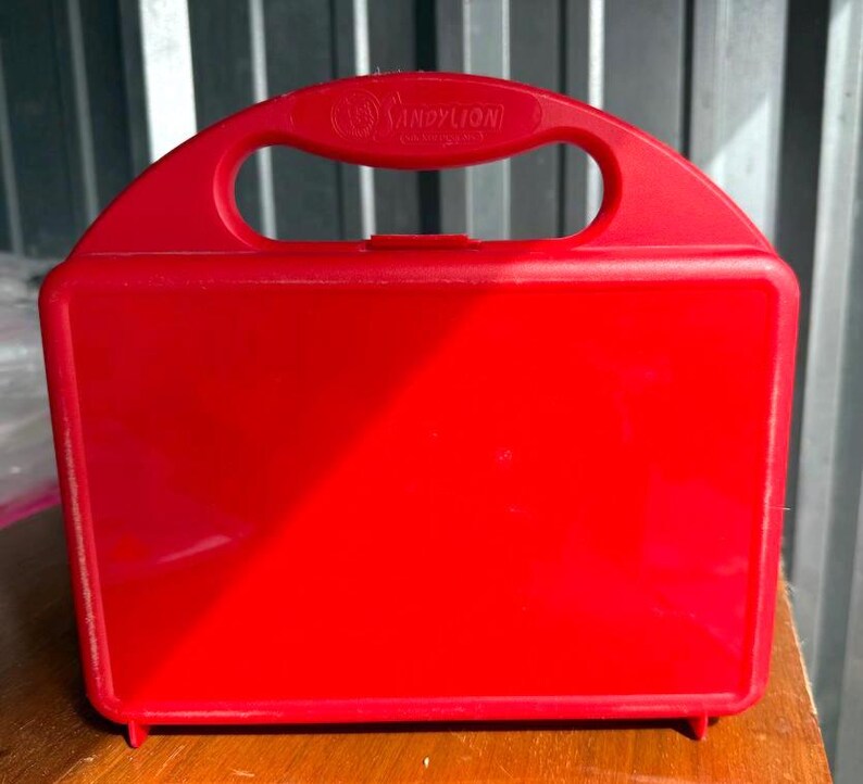 May include: A bright red plastic lunchbox with a handle and the text "SANDY LION" on the handle. The lunchbox is rectangular with rounded corners and sits on a wooden surface.