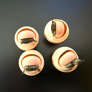 May include: Four small, round, light pink containers with a curved opening, each featuring a set of black eyelashes. The containers are arranged in a square pattern against a dark background.